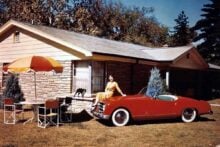 A woman in a yellow dress sits on the hood of a red vintage convertible parked in front of a mid-century house; a black cat walks on a patio table with an umbrella and chairs in the yard.