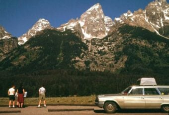 Three people stand near a classic station wagon with a rooftop carrier, admiring tall, snow-dusted mountain peaks under a clear blue sky.