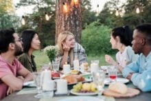 Five people sit outdoors at a table set with food, drinks, and candles. They are talking and smiling, surrounded by greenery and string lights hanging above, suggesting a relaxed, festive gathering.