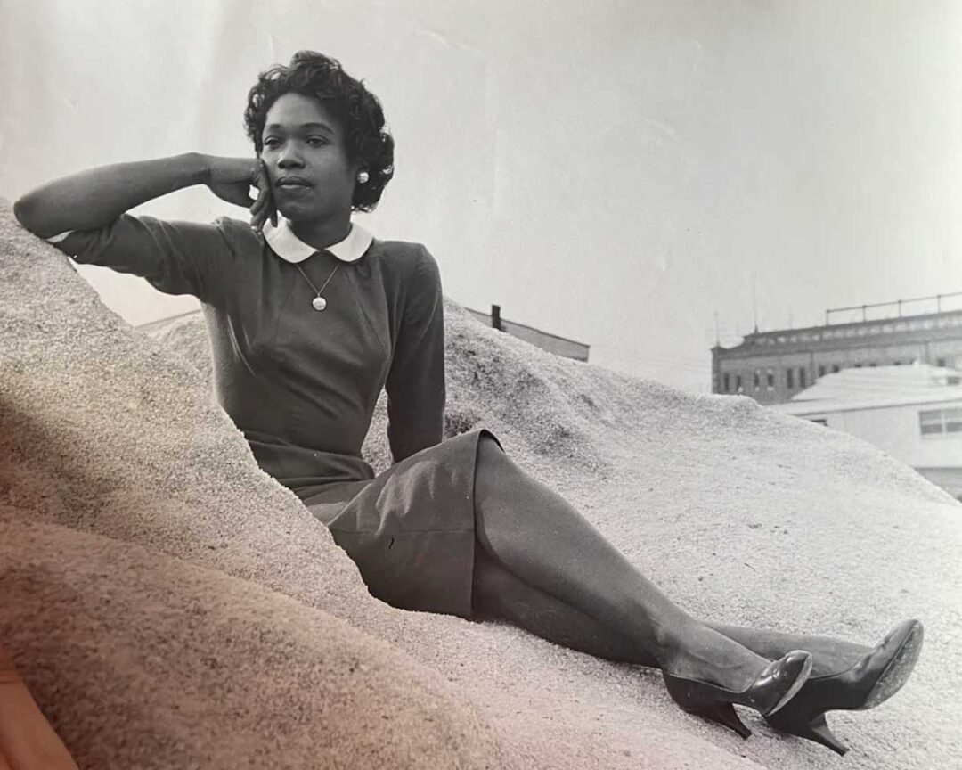 A woman in a mid-century dress and heels sits on a sandy hill, resting her chin on her hand, and gazing thoughtfully into the distance. Buildings are visible in the background. The photo is black and white.