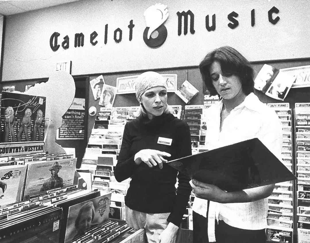 Two women browse vinyl records in a retro music store called Camelot Music. One woman, wearing a headscarf, points at a record while the other holds an album and looks at it. Shelves of records fill the store.