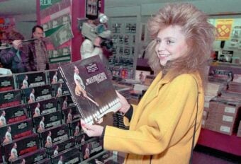 A woman with voluminous hair and a yellow coat smiles while holding a Bruce Springsteen vinyl album in a record store filled with stacked albums and people in the background.