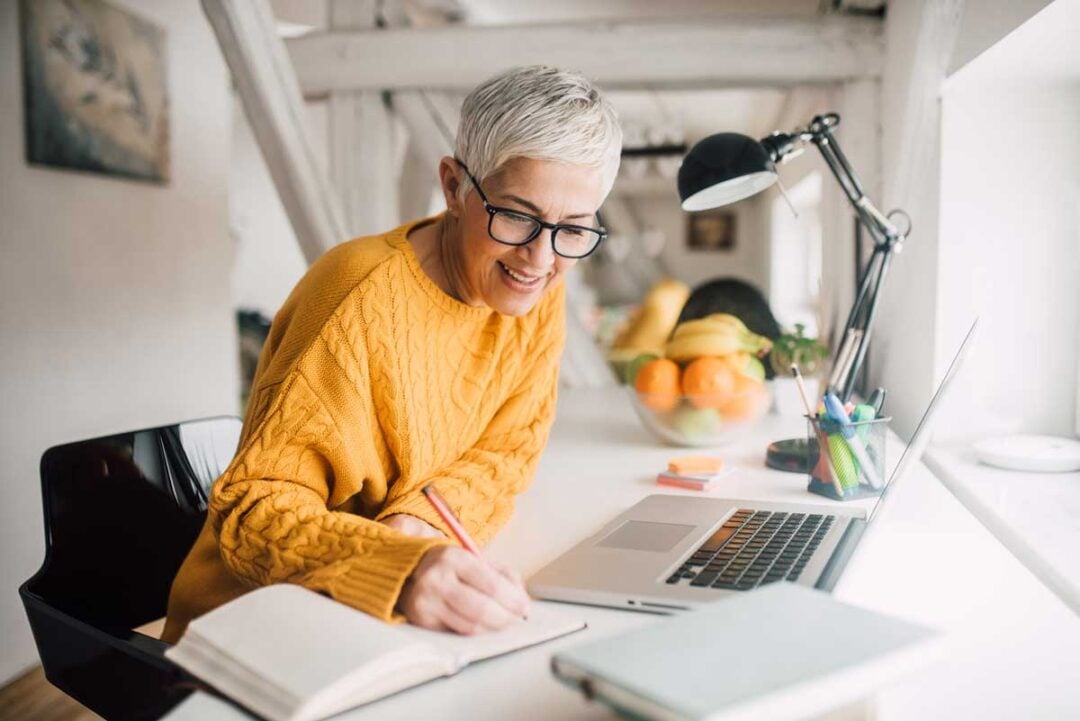 Smiling person with short gray hair and glasses wearing a yellow sweater, sitting at a desk with a notebook, laptop, lamp, and a bowl of fruit, writing while looking at the laptop screen.