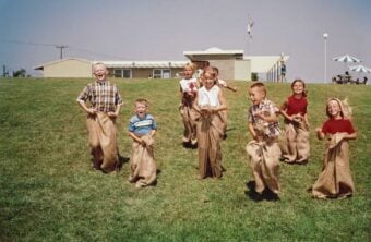 A group of children are laughing and racing in burlap sacks on a grassy hill under a clear blue sky, with a building and umbrellas in the background.