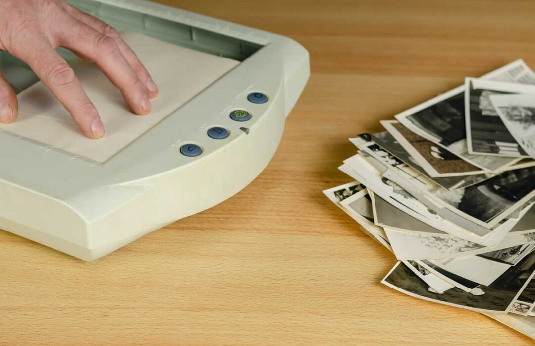 A hand places a photo on a flatbed scanner, with a stack of black-and-white photographs lying on a wooden desk nearby.
