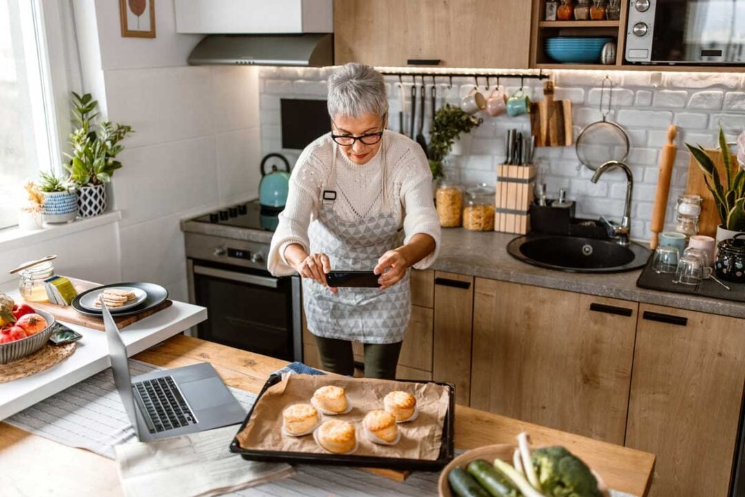 An older woman wearing an apron takes a photo of freshly baked biscuits on a tray in a modern kitchen, with a laptop and various vegetables on the wooden counter nearby.