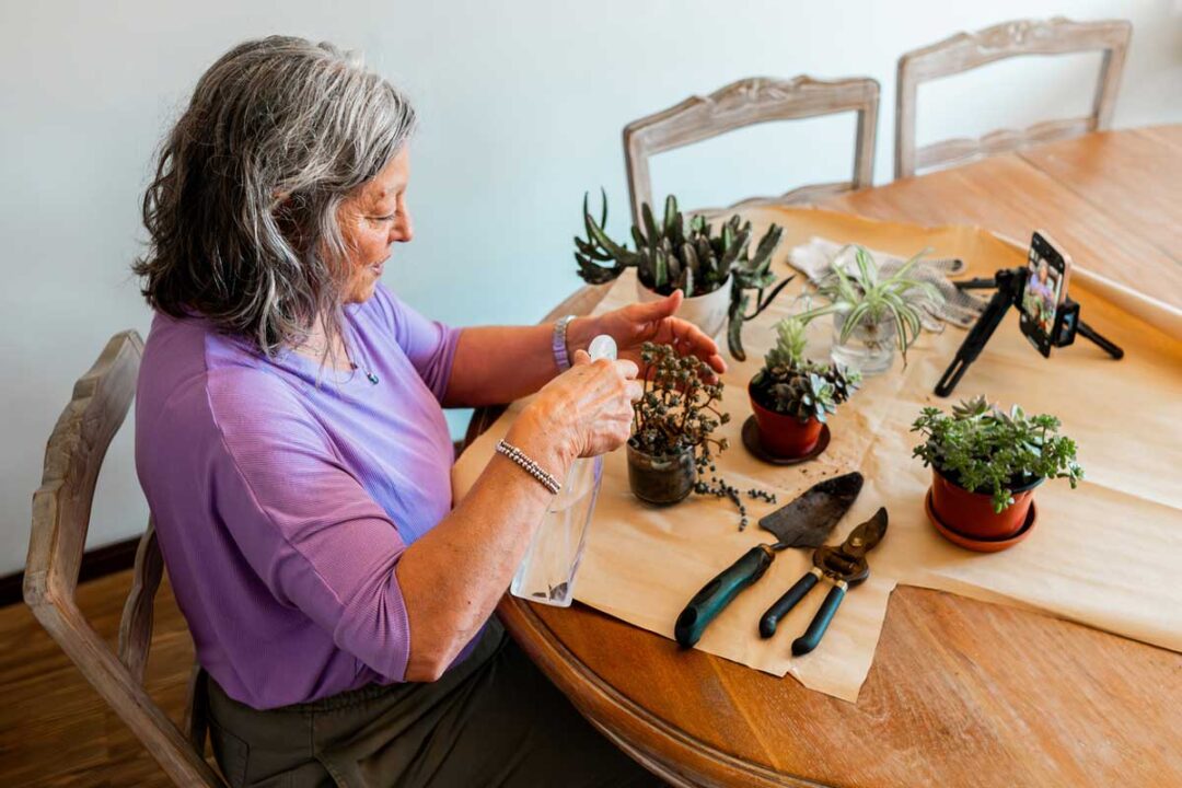 An older woman with gray hair sits at a wooden table tending to potted plants. Gardening tools are laid out, and a smartphone on a tripod records her as she works.