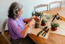 An older woman with gray hair sits at a wooden table tending to potted plants. Gardening tools are laid out, and a smartphone on a tripod records her as she works.