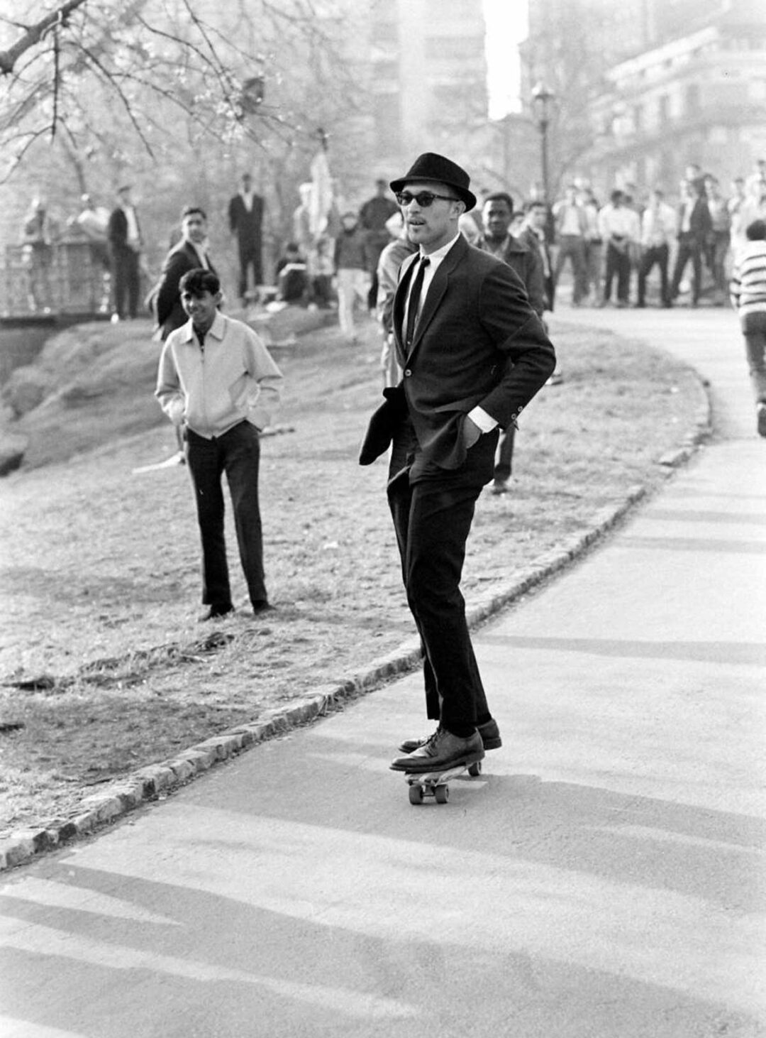 A man in a suit, tie, and hat skateboards on a park path, hands in pockets, while people walk or watch in the background. The scene appears to be from a past decade.