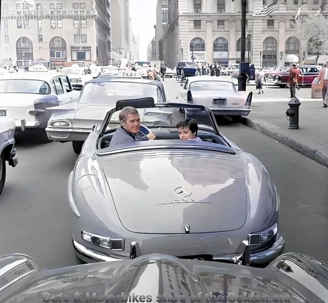A man and a boy sit in a classic silver convertible Mercedes-Benz surrounded by vintage cars and taxis in a busy city street, with tall buildings visible in the background.