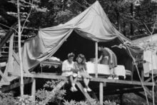Three women at a campsite; two sit on steps reading a book, while one stands in a large canvas tent behind them, surrounded by beds and trees in a wooded area.