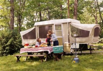 A family enjoys a picnic at a campsite with a pop-up camper in the background. They prepare food on a table covered with a red-and-white checkered cloth in a grassy, wooded area. Camping supplies are visible around them.