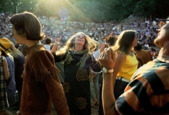 A group of people dance joyfully outdoors at a festival or concert, with sunlight streaming through the trees and a crowd seated in the background on a grassy hillside.