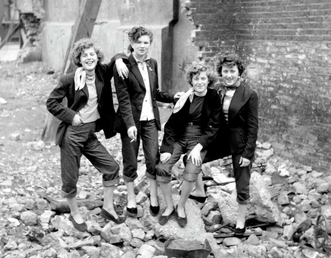 Four young women in suits and scarves stand and sit on rubble in front of a damaged brick wall, smiling confidently. The scene suggests a post-war urban environment.
