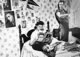 A teenage girl sits on her bed, smiling and holding a James Dean magazine, surrounded by posters and a life-size cutout of James Dean on her wall in a 1950s-themed bedroom.