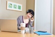 A person wearing headphones sits at a desk with a laptop, coffee cup, pens, notepad, and phone, writing while listening to music. There is artwork on the beige wall behind them.
