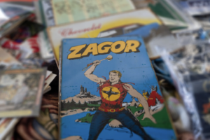 A vintage comic book titled "Zagor" features a muscular man in a red shirt with a yellow emblem, holding an axe, with a castle and forest in the background. The comic is surrounded by other assorted papers.