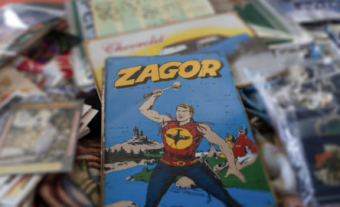 A vintage comic book titled "Zagor" features a muscular man in a red shirt with a yellow emblem, holding an axe, with a castle and forest in the background. The comic is surrounded by other assorted papers.