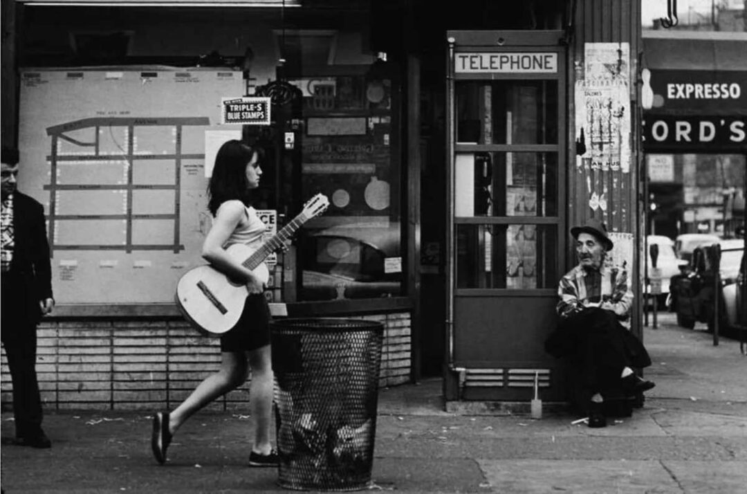 A woman carrying a guitar walks past a trash can on a city sidewalk. An older man wearing a hat sits near a phone booth, watching her. The background shows storefronts and a street scene.