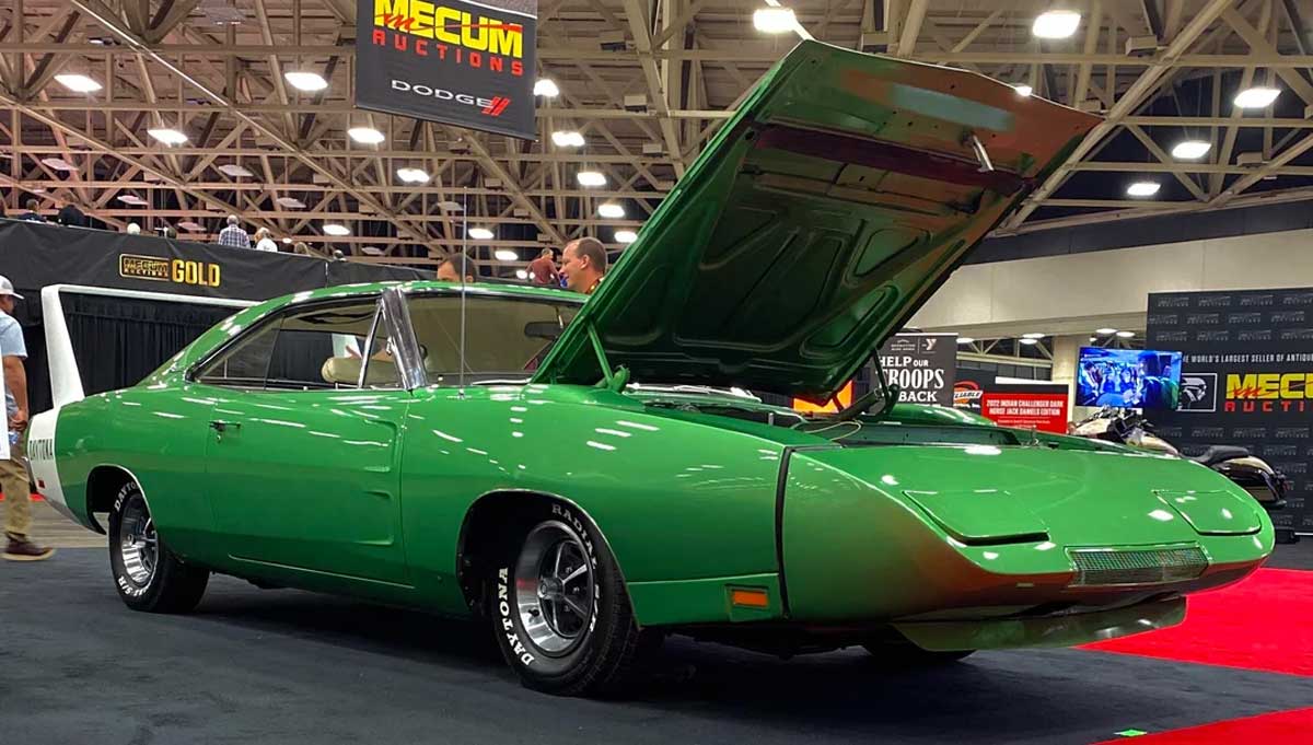 A bright green classic Dodge Charger Daytona with its hood open is displayed at an indoor car auction, under bright lights and surrounded by banners and other vehicles.