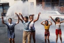 Five children laugh and play in the street, arms raised, as water sprays from an open fire hydrant behind them on a sunny day. A parked car and storefronts are visible in the background.