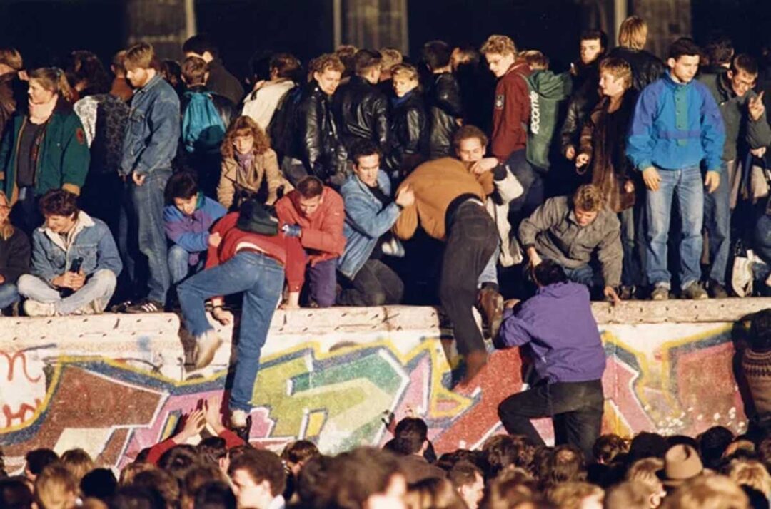 A large crowd gathers at the Berlin Wall, with people climbing over and sitting on the graffiti-covered barrier, celebrating its fall at night.