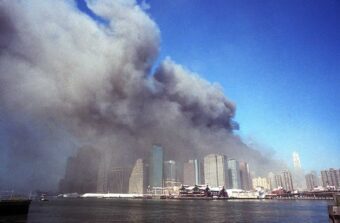 Thick, dark smoke billows from several skyscrapers in a city skyline under a clear blue sky, with water and docks in the foreground.