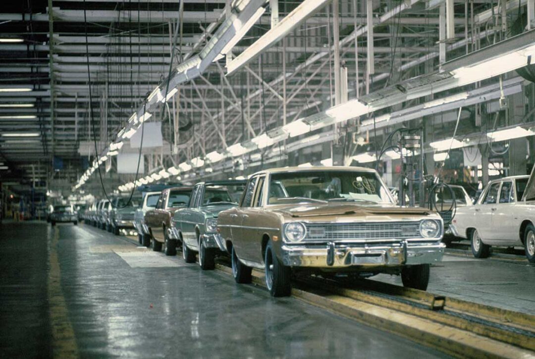 A row of vintage cars moves along an automobile assembly line in a large, brightly-lit factory with overhead lights and metal beams.