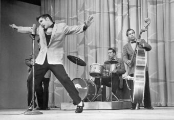 A lively performer strikes a dramatic pose at a microphone while a drummer and a double bass player accompany him on stage; the backdrop features draped curtains.
