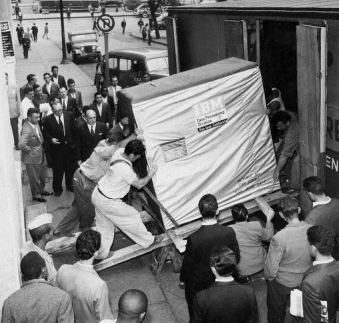 A group of men work together to move a large IBM Data Processing System into a building using a ramp, as a crowd gathers on a busy city sidewalk to watch. The scene is in black and white and appears historical.