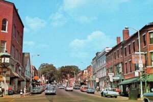 A busy small-town street in the 1960s or 1970s, lined with brick buildings, vintage cars parked along the curb, storefronts with colorful signs, and people walking on the sidewalks under a bright blue sky.
