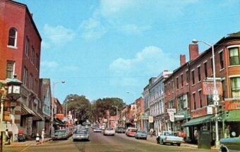 A busy small-town street in the 1960s or 1970s, lined with brick buildings, vintage cars parked along the curb, storefronts with colorful signs, and people walking on the sidewalks under a bright blue sky.