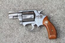 A silver revolver with a short barrel and a wooden grip lies on a textured gray surface.