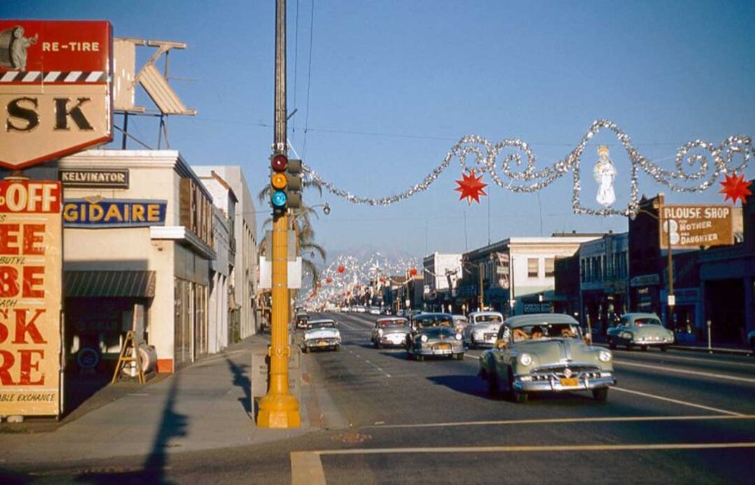 A 1950s city street decorated with Christmas garlands and red stars; vintage cars drive by, and storefronts with colorful signage line both sides of the street under a clear blue sky.