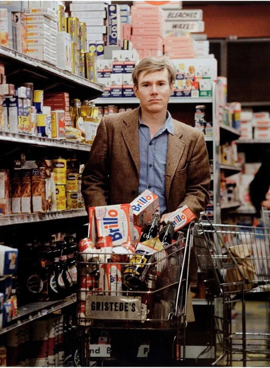 A man in a brown blazer and blue shirt stands in a grocery aisle, holding a shopping cart filled with Brillo boxes and various groceries. Shelves stocked with food products line both sides of the aisle.