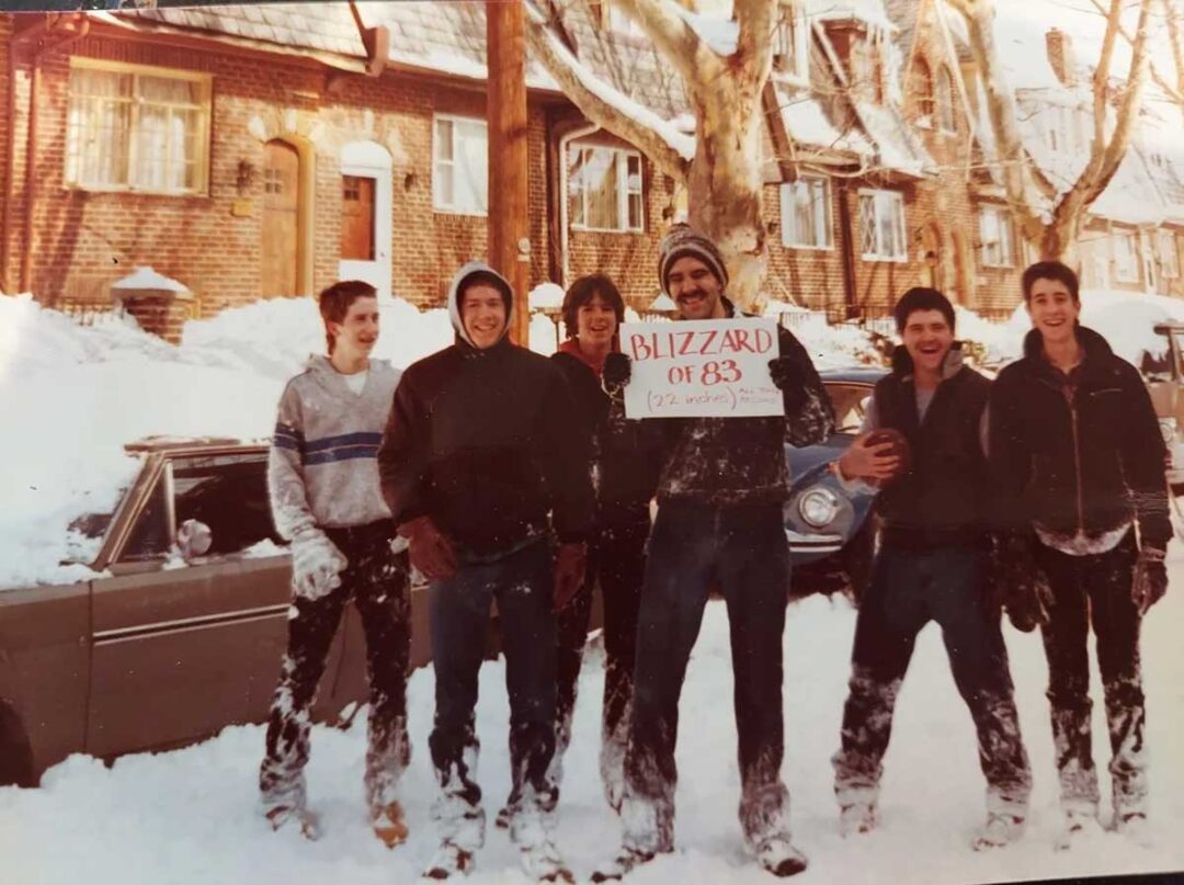 Six smiling people in winter clothes stand in deep snow beside a car and houses. One holds a sign reading "BLIZZARD OF '83 (27 inches)." Snow covers the ground and the parked cars.