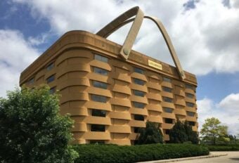 A large building designed to look like a woven picnic basket, complete with two giant handles on the roof, sits surrounded by trees and greenery under a partly cloudy sky.