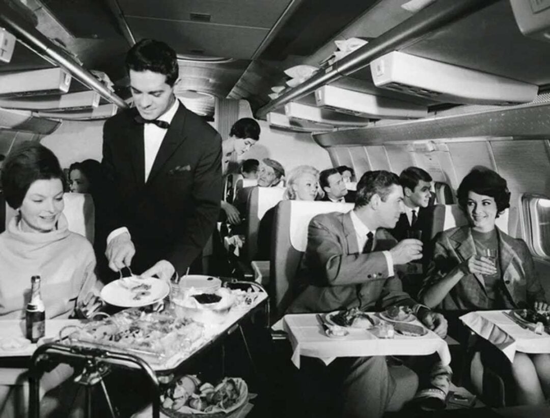 A black-and-white photo of passengers in an airplane cabin, seated comfortably and enjoying a meal. A flight attendant in a suit serves food from a cart to a smiling woman. The scene depicts a vintage, luxurious in-flight dining experience.