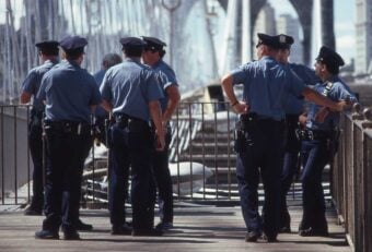 A group of police officers in blue uniforms stand and talk together on a bridge during the daytime. The bridge's structure and cables are visible in the background.