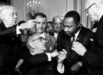 A group of men, including Martin Luther King Jr., gather closely as one man seated in the center hands a pen to King, surrounded by conversation and attention in a formal setting.