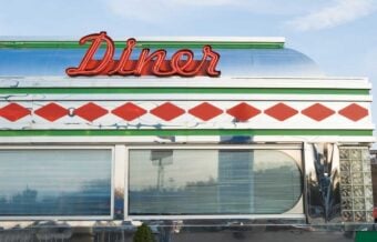 A retro-style diner with a shiny metal exterior, red diamond patterns, and a large red neon "Diner" sign on the roof. The building has horizontal windows with closed blinds.