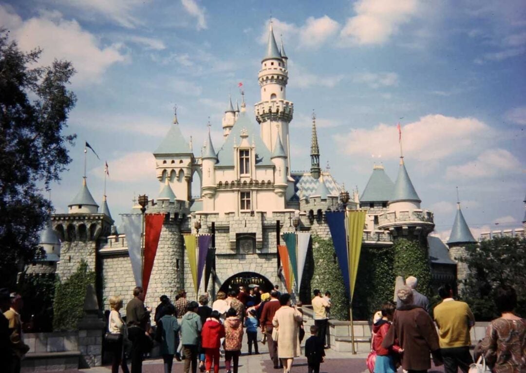 A group of people walk toward Sleeping Beauty Castle at Disneyland on a sunny day, with colorful banners lining the path and blue sky with clouds above the castle.