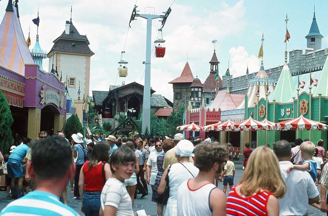 A busy amusement park scene with crowds walking between colorful, castle-like buildings. Overhead, cable cars move along a skyway. The day is bright and sunny, and people are dressed in summer clothes.