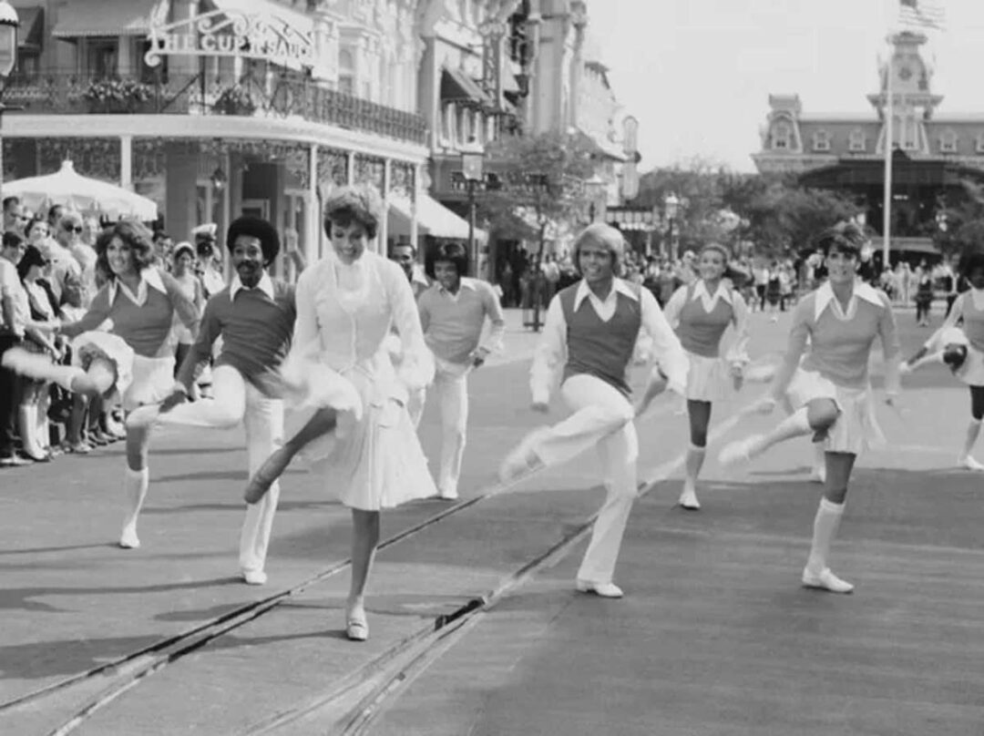 A group of performers, including a woman in a dress and people in matching outfits, dance in formation on a street in front of a crowd at a theme park, with buildings and a railroad station visible in the background.