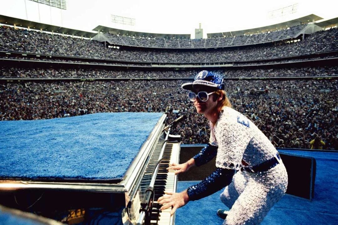 A performer wearing a sequined outfit, cap, and large sunglasses plays the piano on a stage, facing a massive, cheering crowd in a stadium.