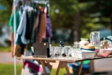 A table at a yard sale displays glassware, bowls, a radio, and various household items. In the background, clothing hangs on a rack with green trees and sunlight visible.