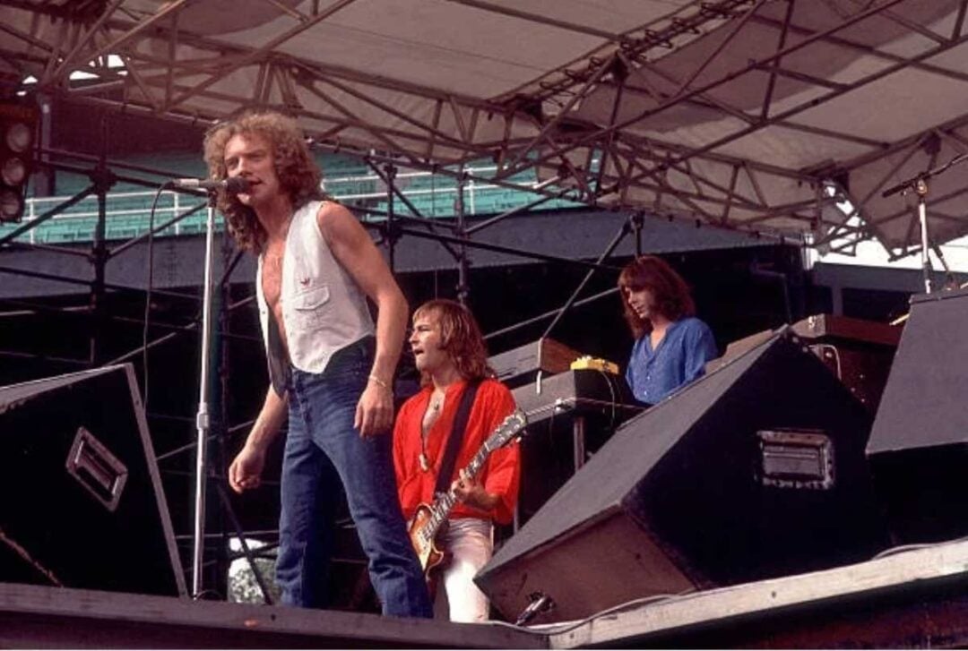 A rock band performs on an outdoor stage. The lead singer stands at a microphone, while two musicians, one with a guitar and one at a keyboard, play behind him. The stage is covered by a metal canopy.