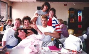 A group of six young adults pose and smile together in a casual cafeteria setting, with colorful chairs, a table with bags and drinks, and a vending machine in the background.