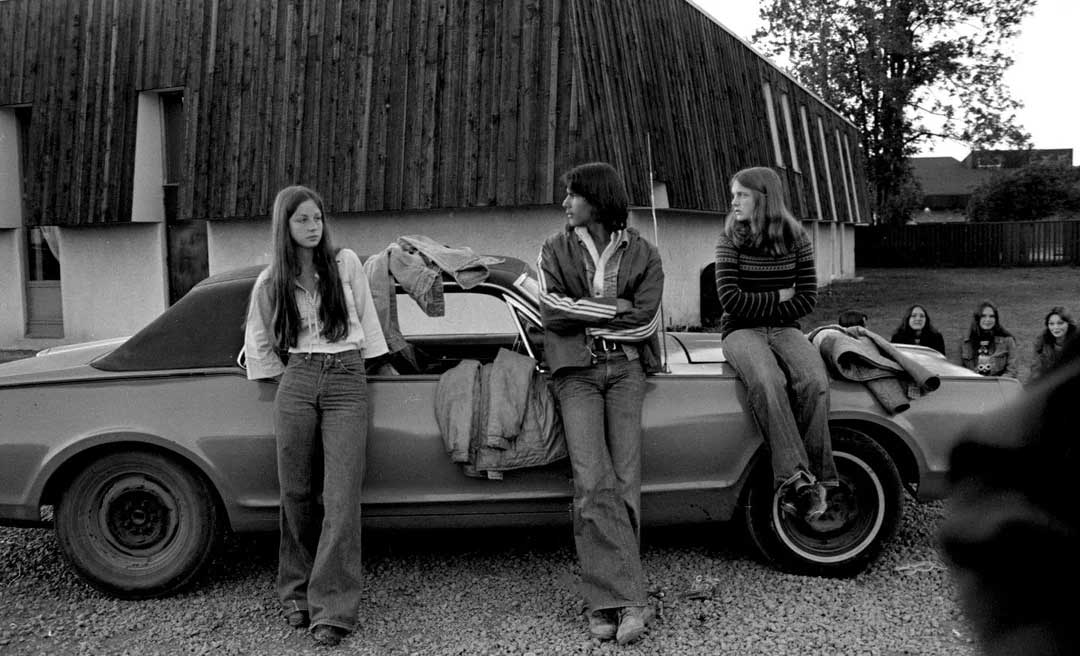Three teenagers in 1970s clothing lean against a classic car parked outside a wooden building, chatting casually. In the background, a group of people sit on the ground near the building.
