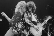 Two rock musicians perform energetically on stage; one sings into a microphone while the other plays an electric guitar. Both have long, curly hair and wear patterned shirts, with stage lighting and a drum set visible in the background.